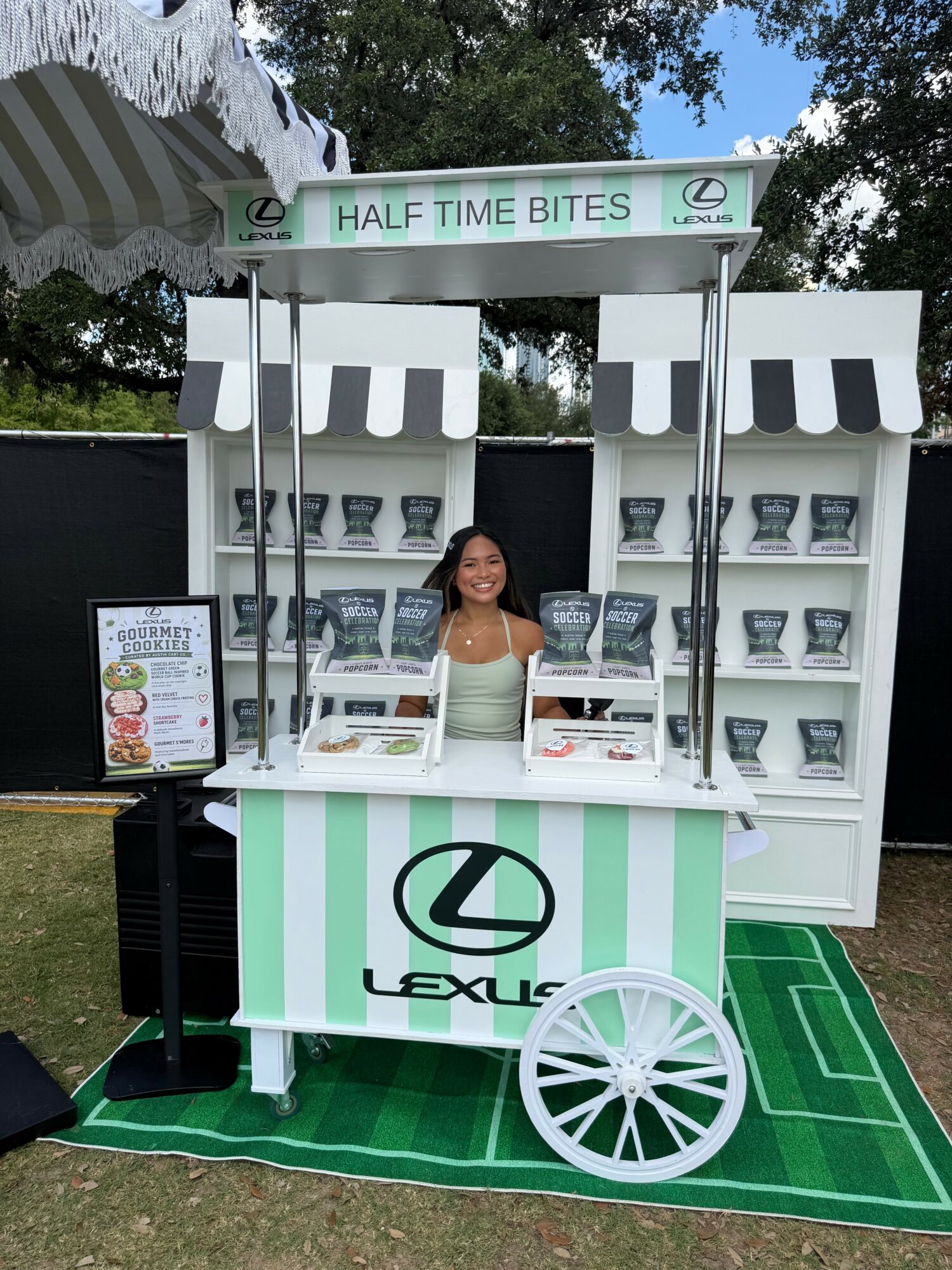 Young woman standing behind a lemonade stand with shelves of cups, a sign reading 'Half Time Bites,' and a green mat.