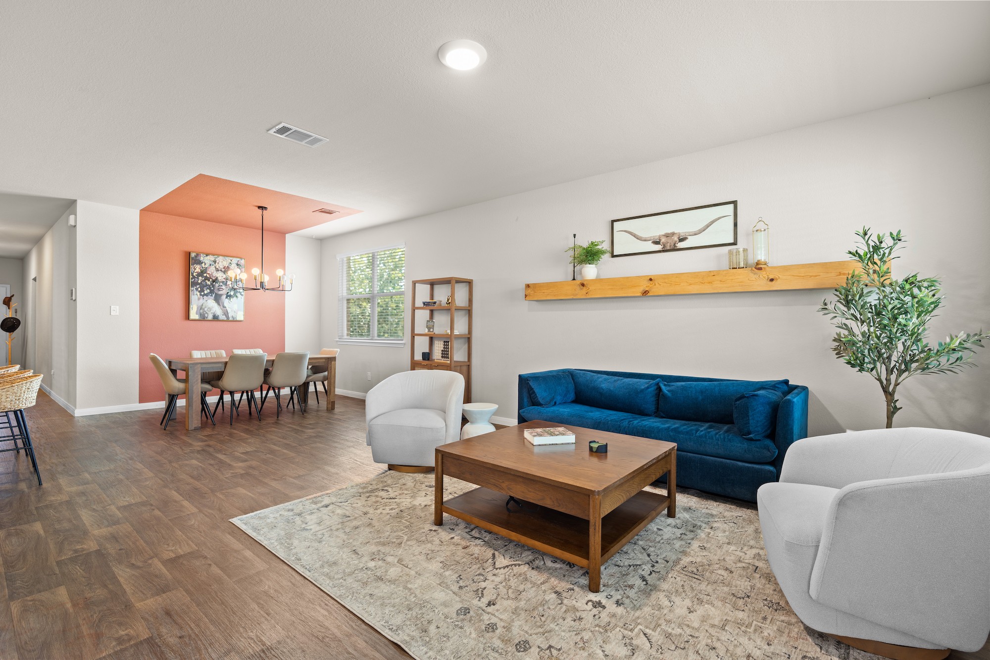 Living room with blue sofa, white armchairs, wooden coffee table, and a wall shelf with decor, adjacent to dining area with pink accent wall.