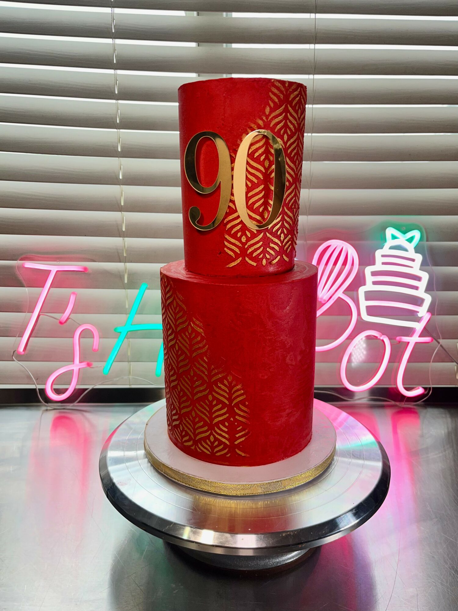 Two red cylindrical cakes with gold designs and numbers, on a silver cake stand, in front of a window with blinds.