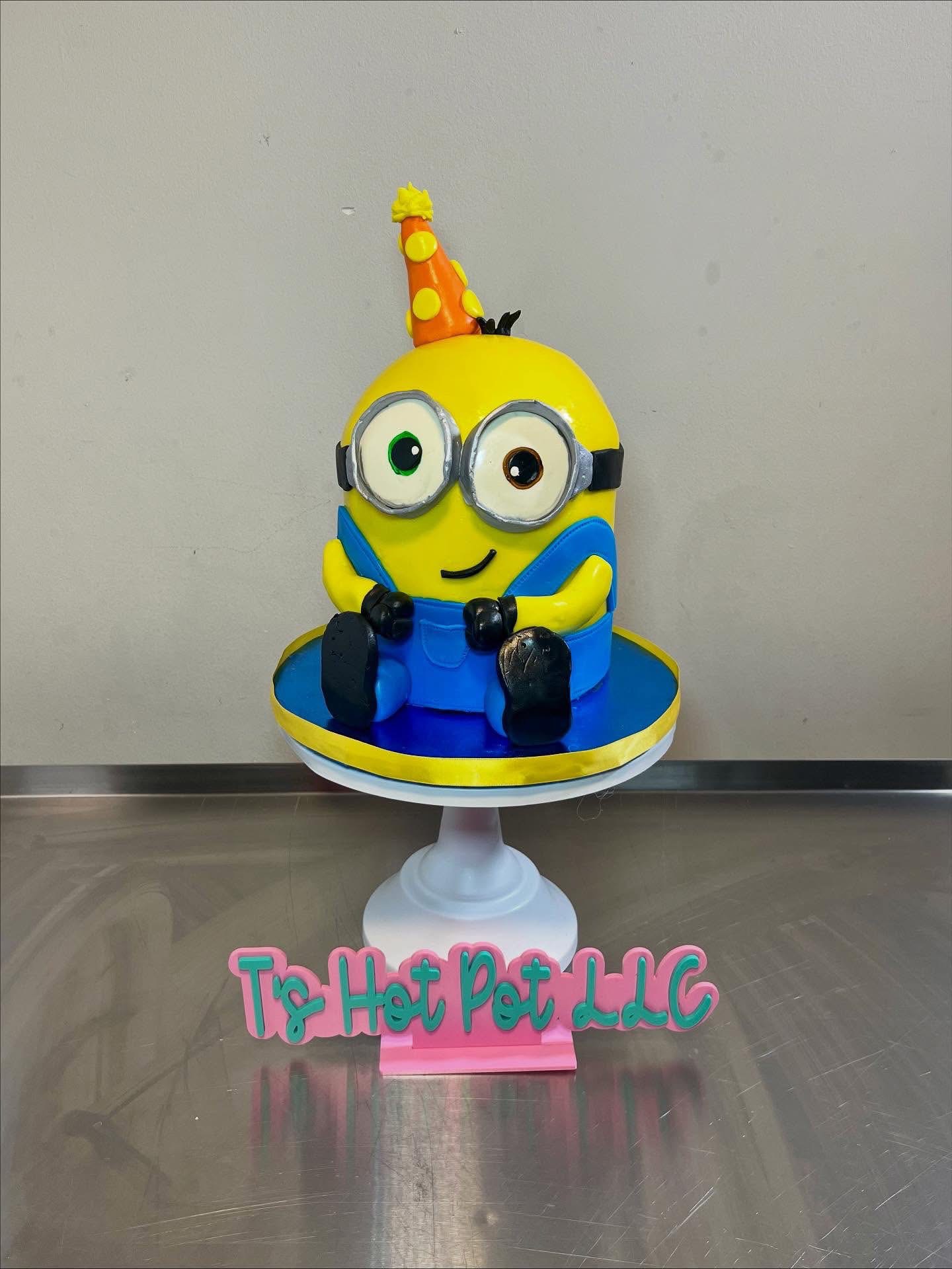 Minion cake with a party hat, sitting on a white cake stand, with a pink sign reading 'Tip Hat Pot LLC' in front.