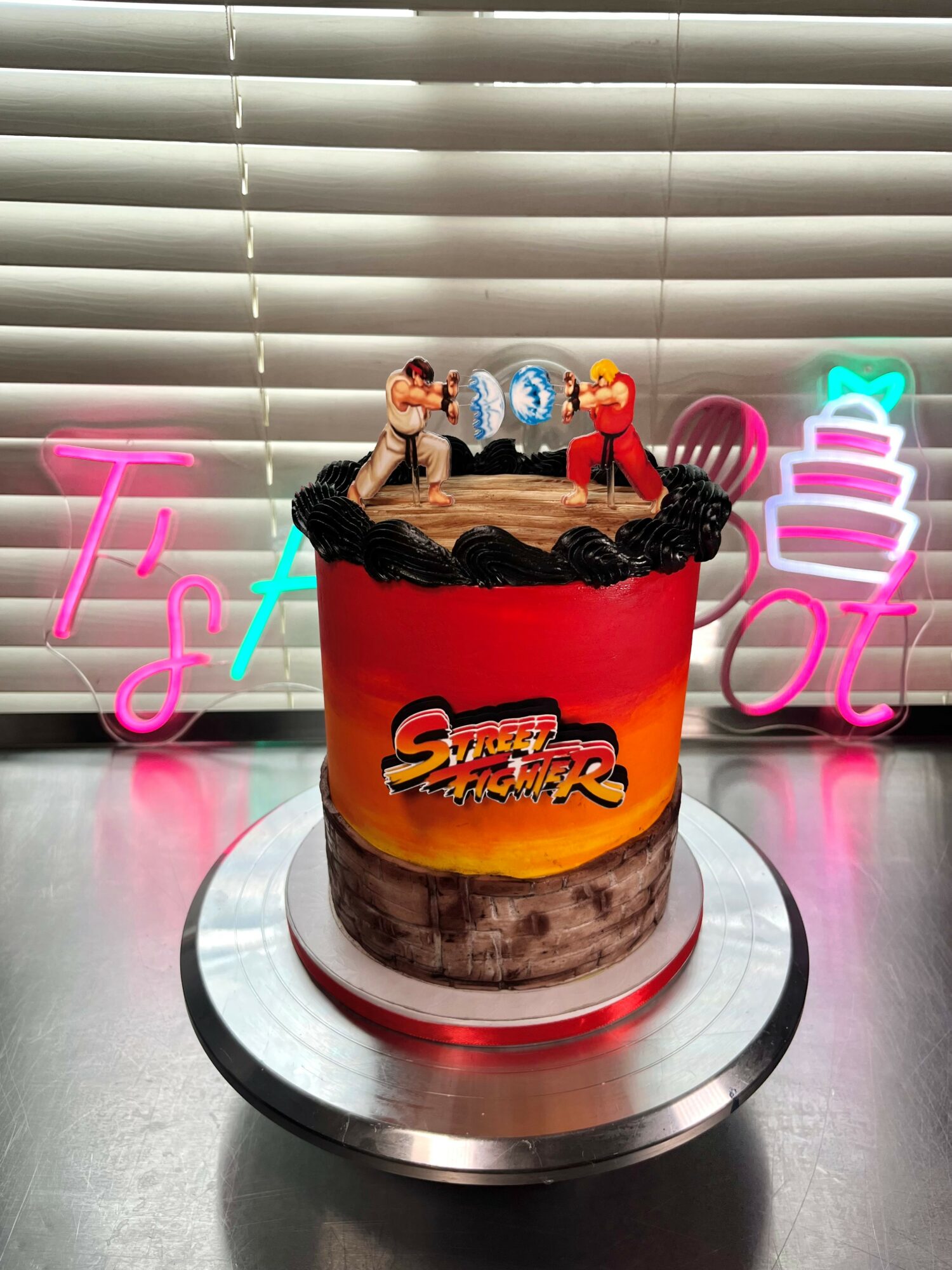 Decorative cake with figurines on top, colorful neon signs in the background, and a silver cake stand.