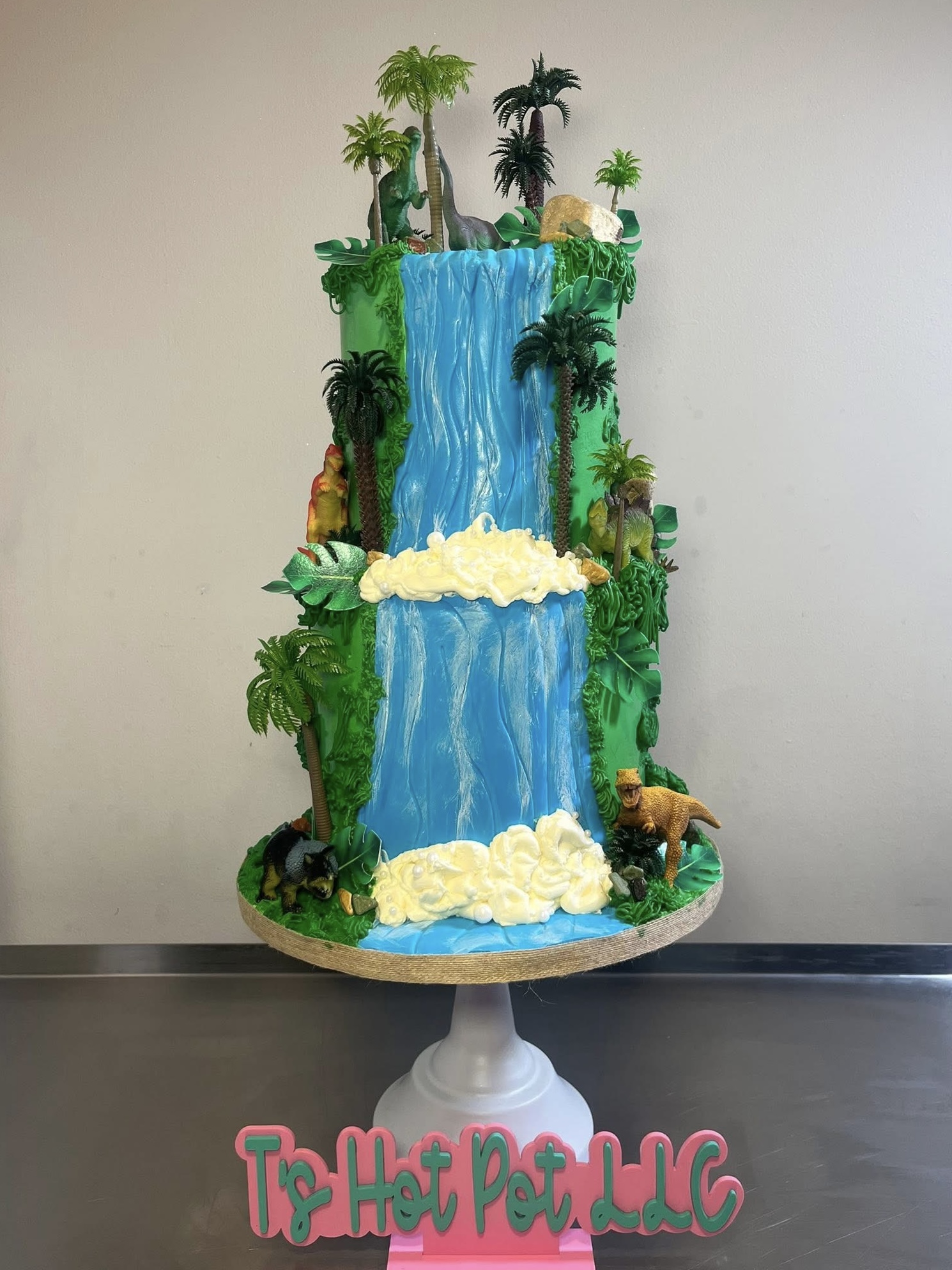 Three-tiered cake with waterfalls, trees, and animals, decorated with green foliage and white clouds, on a white stand.