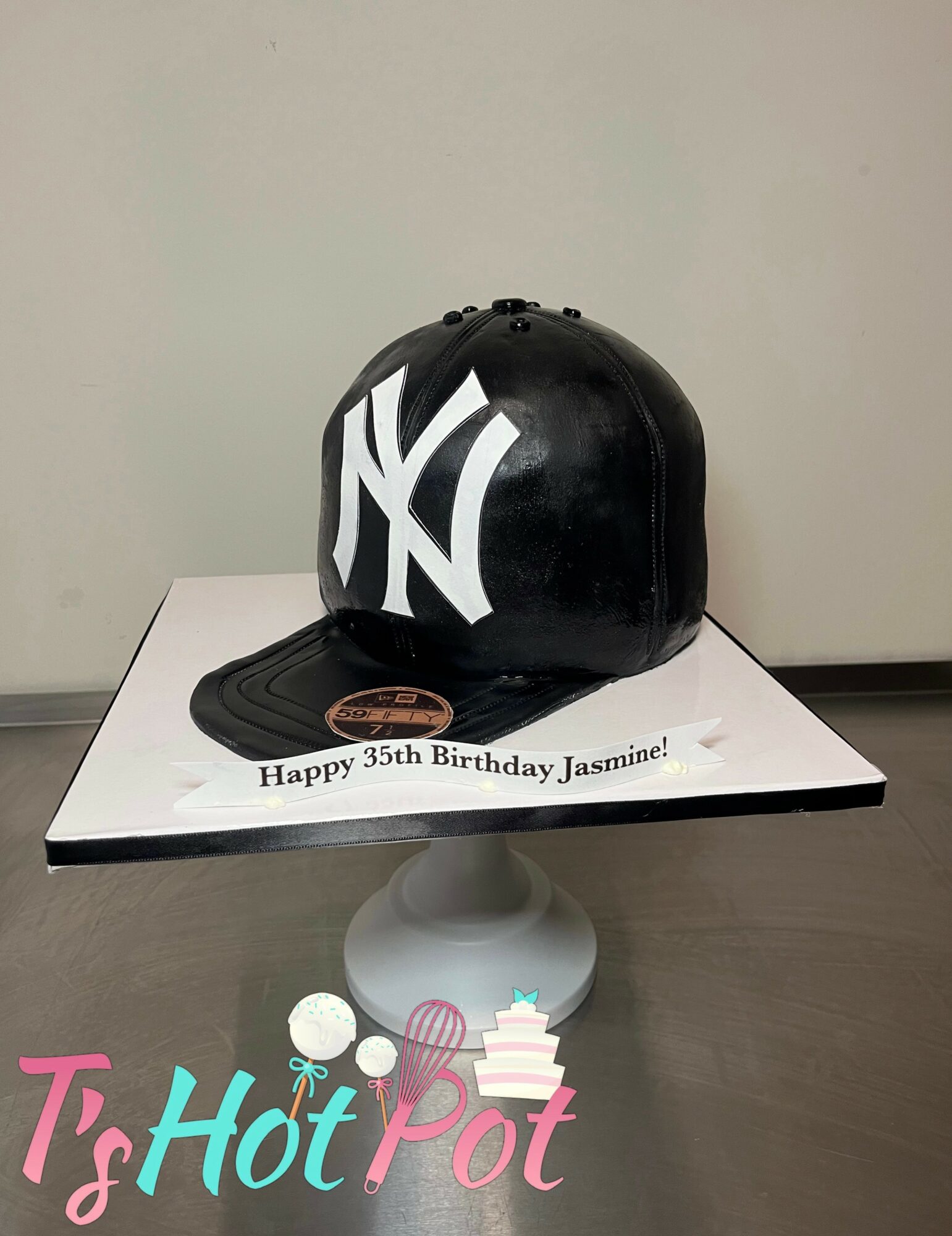 Black baseball cap with white New York Yankees logo on a cake, celebrating Jasmine's 35th birthday, on a white cake stand.