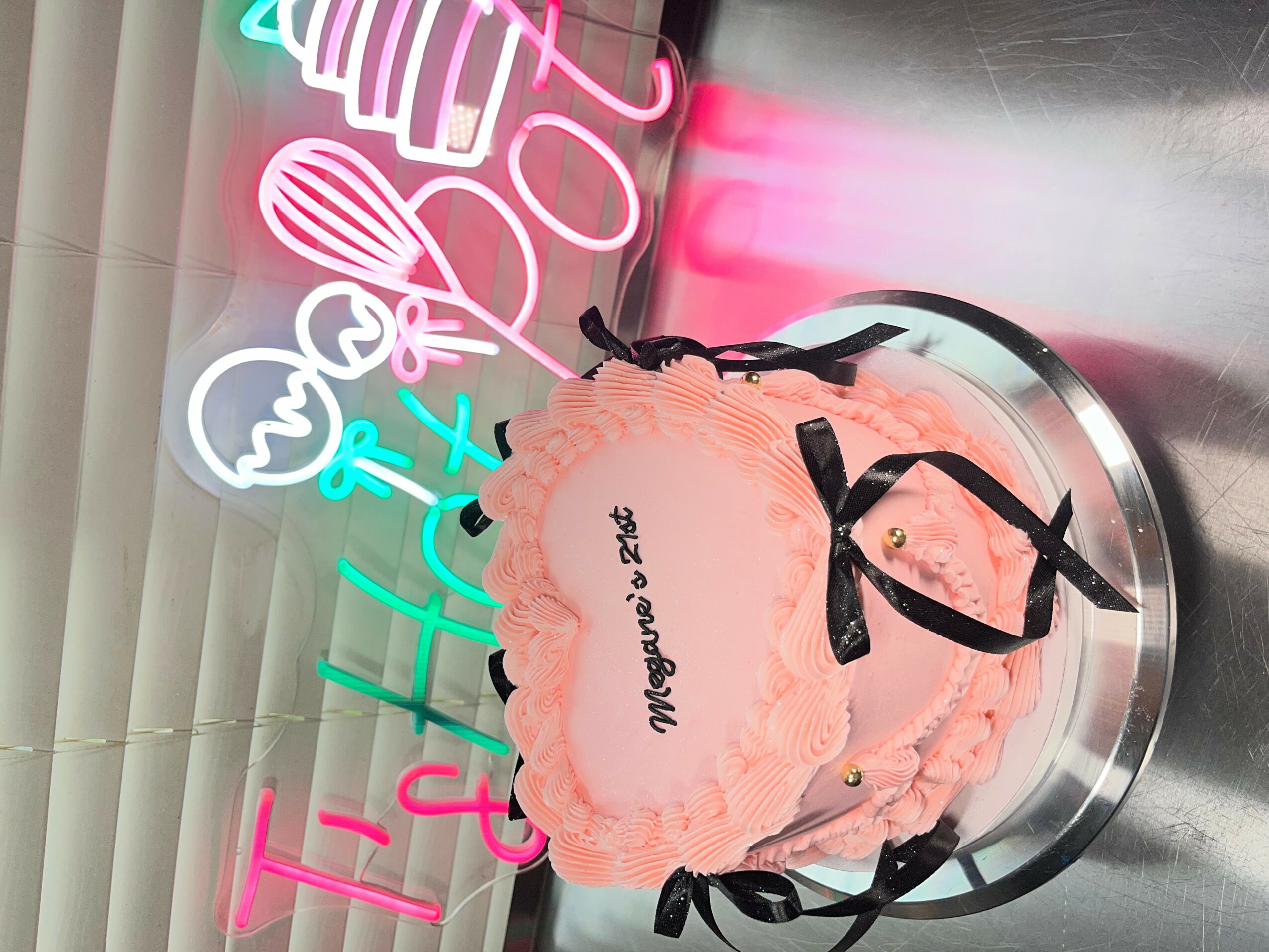 Pink heart-shaped pillow with black ribbon, text, and a small gold charm, placed on a reflective surface.