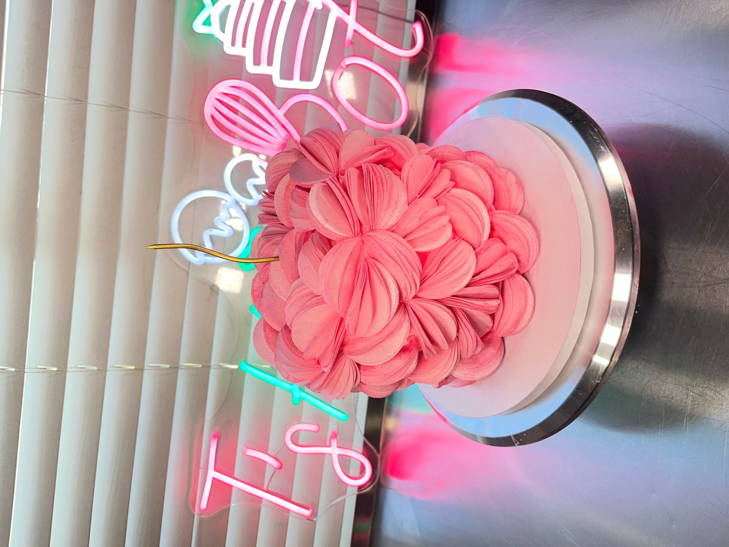 Decorative pink cake with ruffled frosting on a silver stand, neon signs in background, vertical blinds behind.