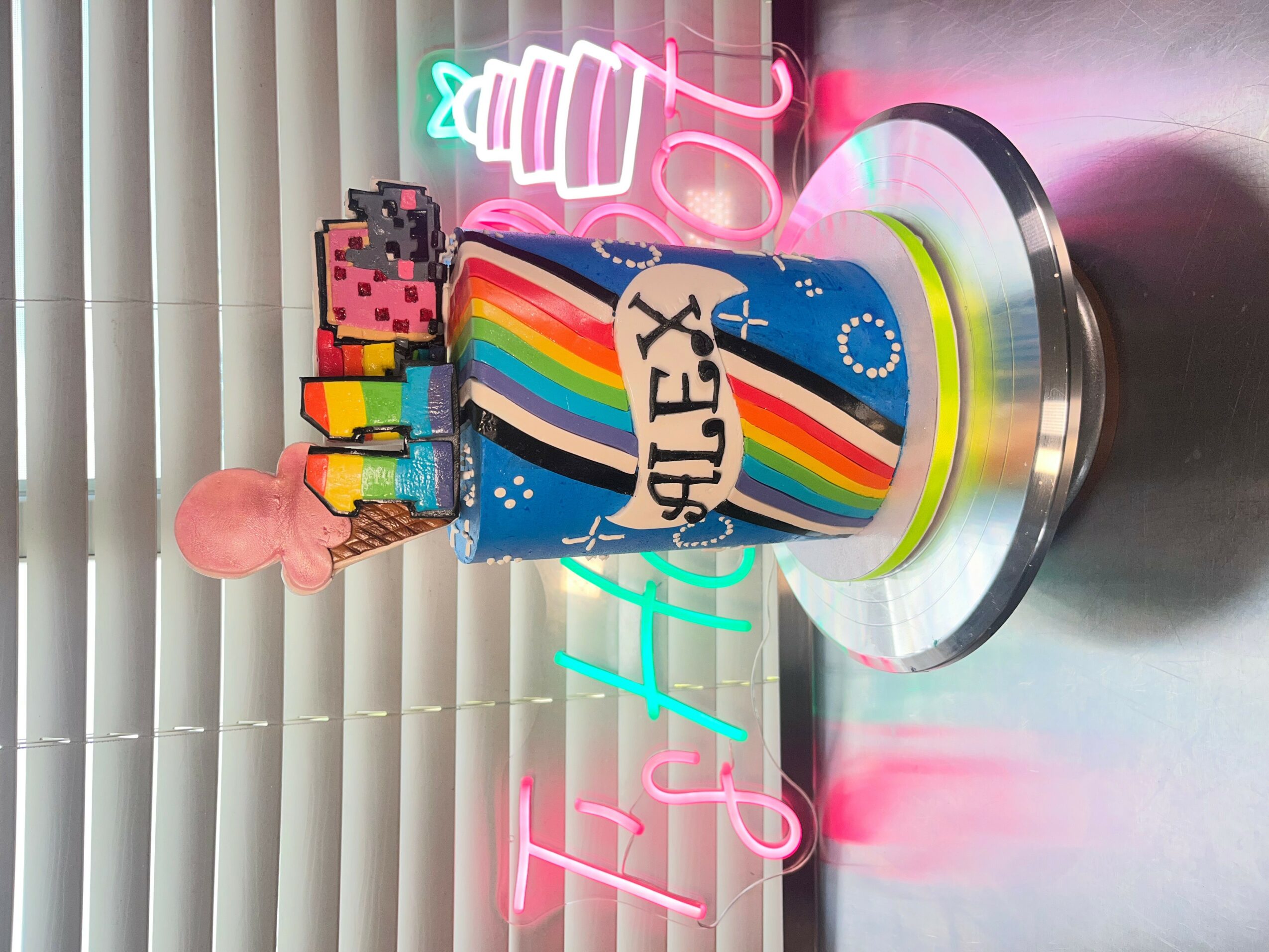 Cake with a rainbow design, pixelated figures, a pink figure, and a neon sign, on a silver base.
