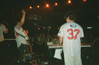 Person with curly hair plays keyboard on stage, others in jerseys, audience in background, dim lighting, concert setting.