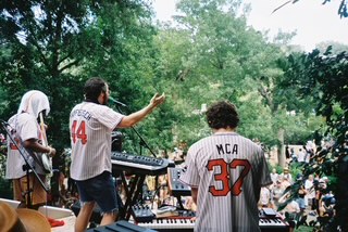 Group of people performing music outdoors with trees in the background, some wearing sports jerseys, one playing keyboard.