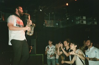 A man playing saxophone on stage with an audience watching, some taking photos, in a dark venue.
