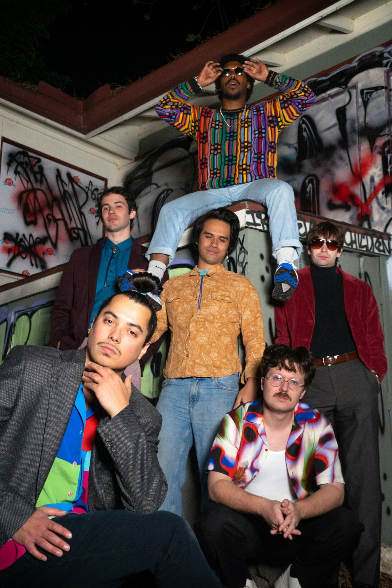 Group of six young people posing in front of graffiti-covered wall, some sitting and some standing, with one person on others' shoulders.