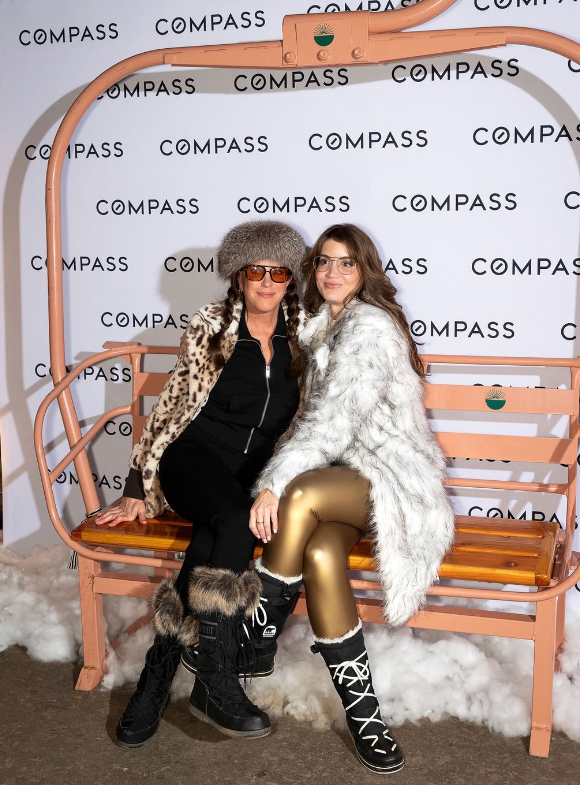 Two women sit on a pink bench with a white backdrop, wearing fashionable winter clothing and accessories.