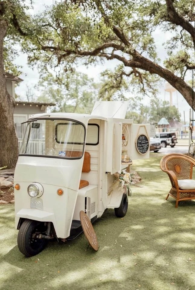 Small white vehicle with a door open, parked on grass under trees, with a wicker chair nearby.