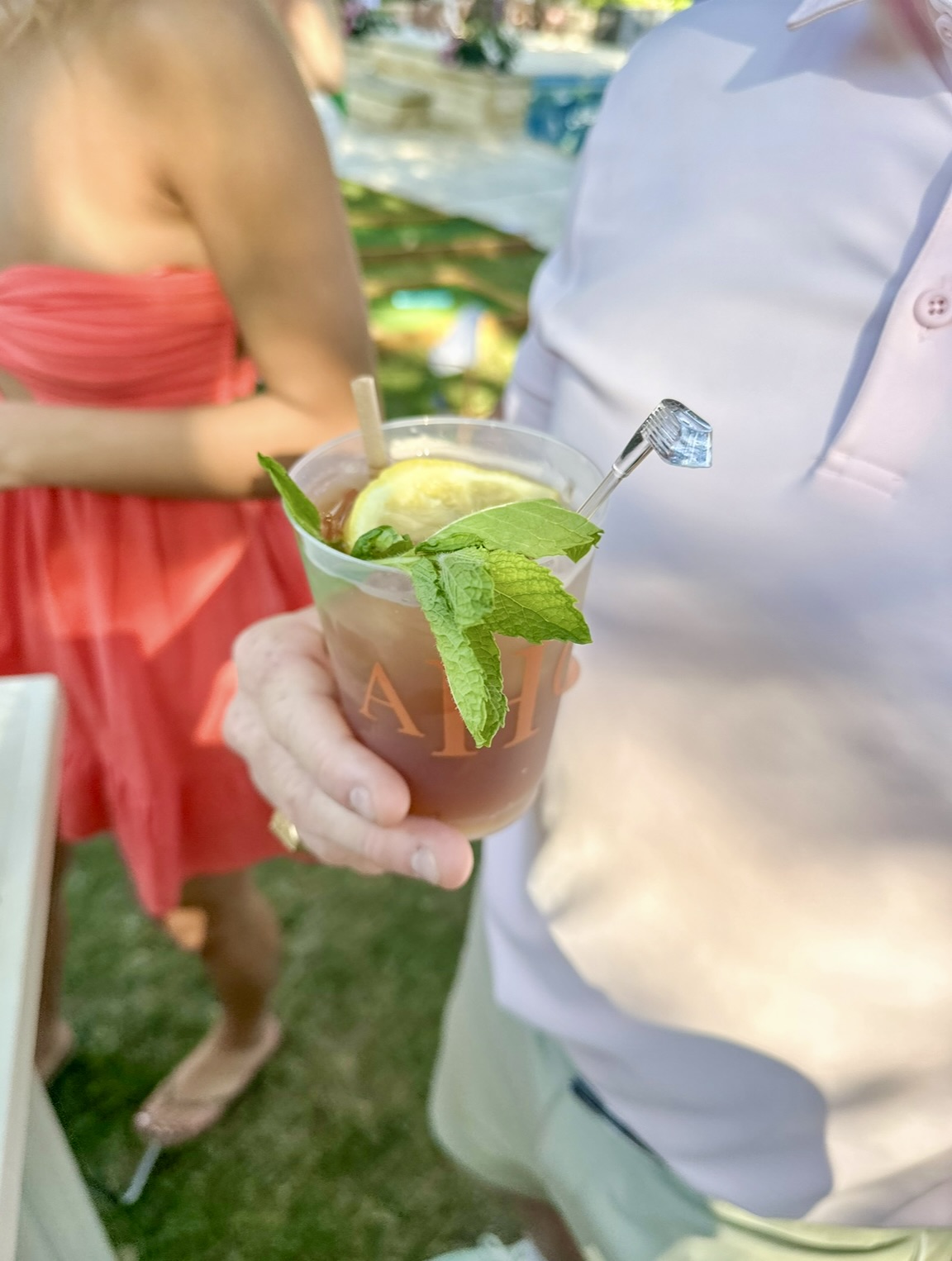 Person holding a cocktail garnished with mint and lemon, outdoors with people in the background.