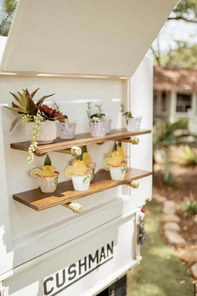 Decorative display with small potted plants and cupcakes on wooden shelves, outdoors with blurred background of trees and buildings.
