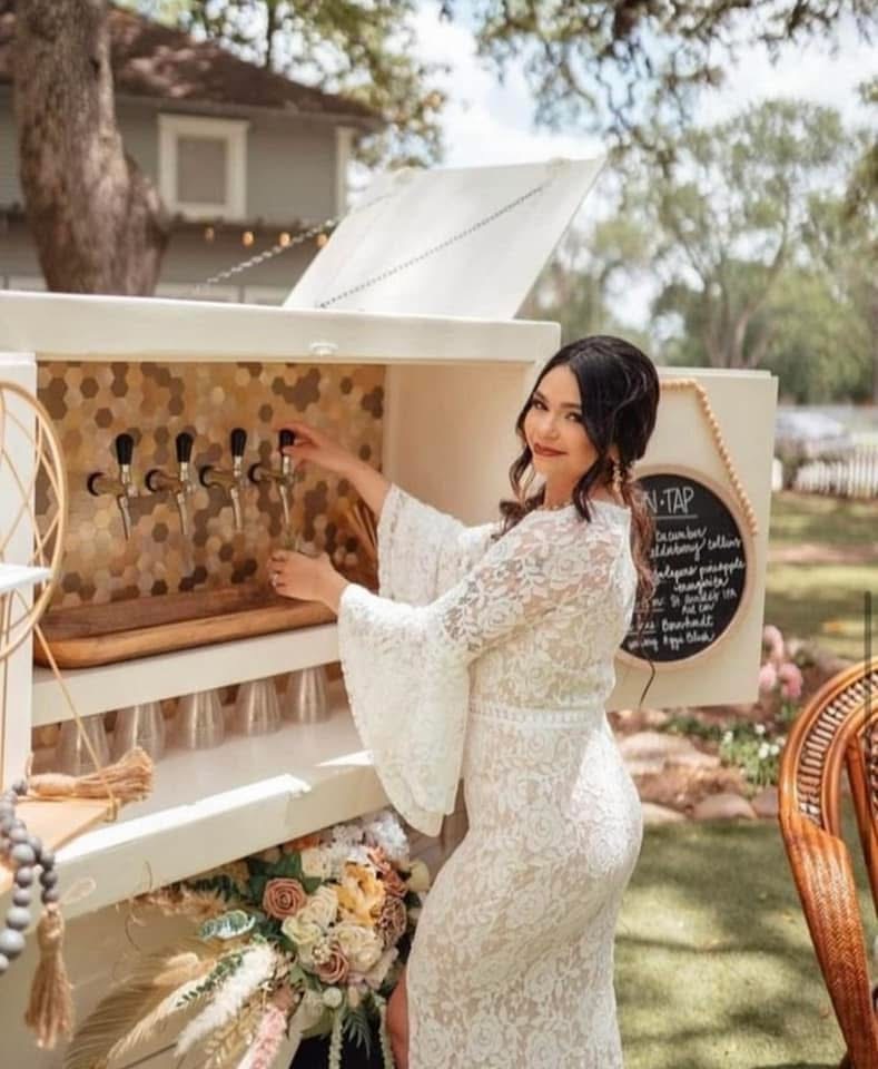 Woman in white lace dress preparing drinks at outdoor bar with floral arrangement, trees, and a chalkboard menu in background.