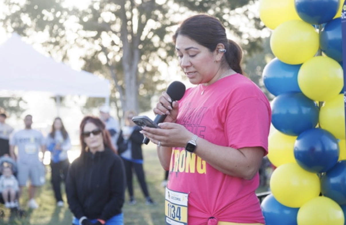 Woman in pink shirt holding a microphone and reading from a phone outdoors, with balloons and people in the background.