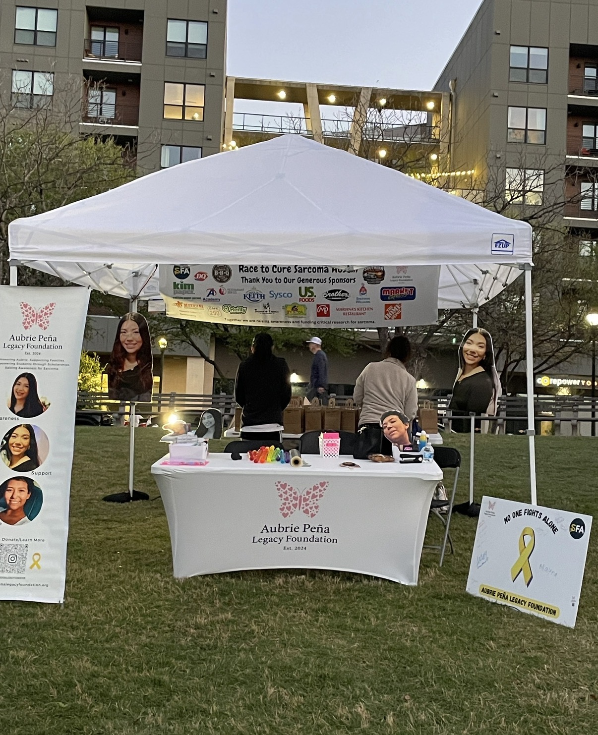 Outdoor booth with a white canopy tent, table with pink and white items, and people in the background, in a park setting.