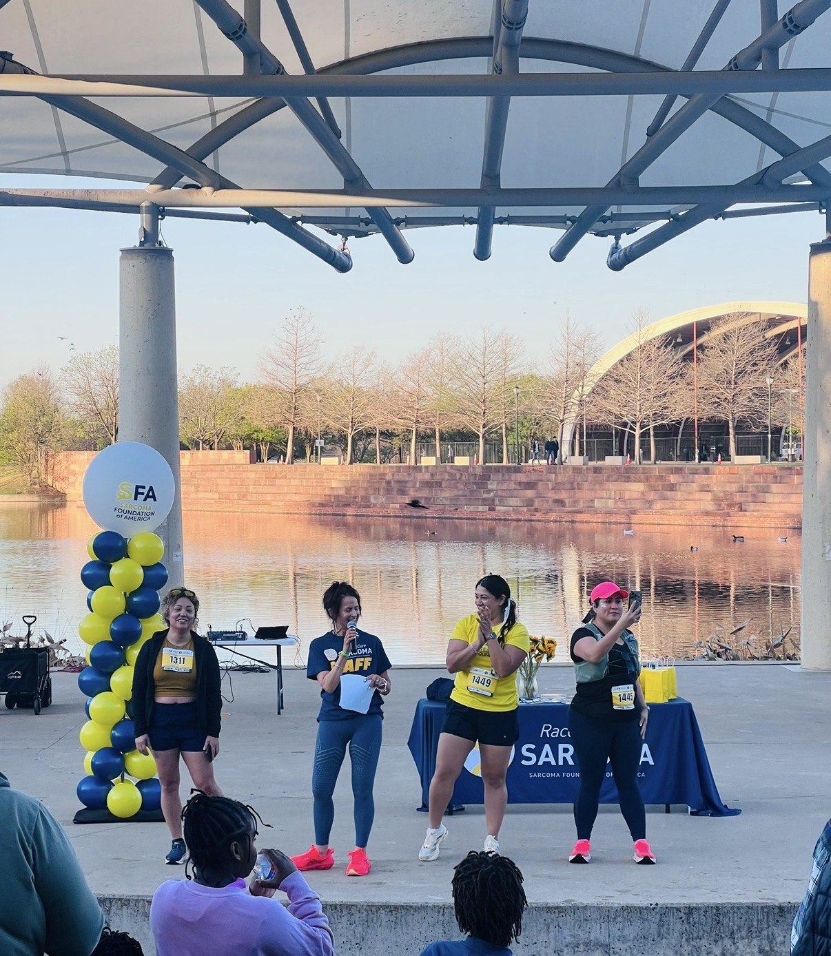 Four women on stage near a river, with a table and balloons, participating in an outdoor event during daytime.