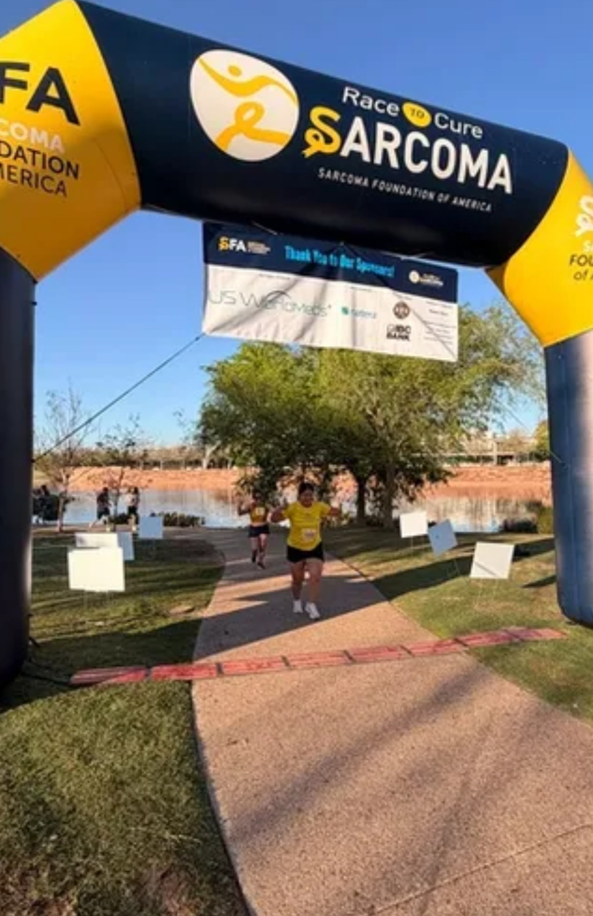 Runner crossing finish line under ARCOMA race arch with trees and blue sky in background.