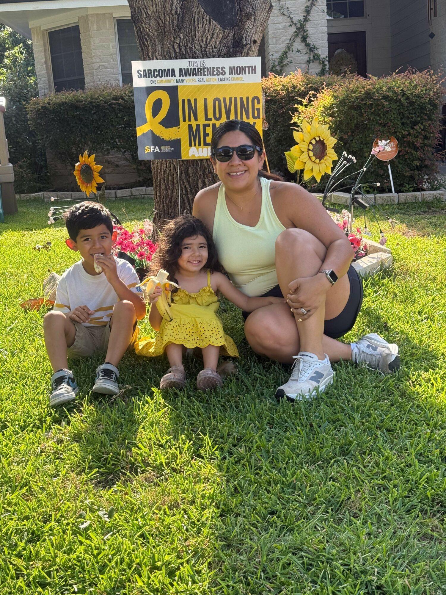 Two women and two children sitting on grass near a sign and flowers, sunny day, greenery around.