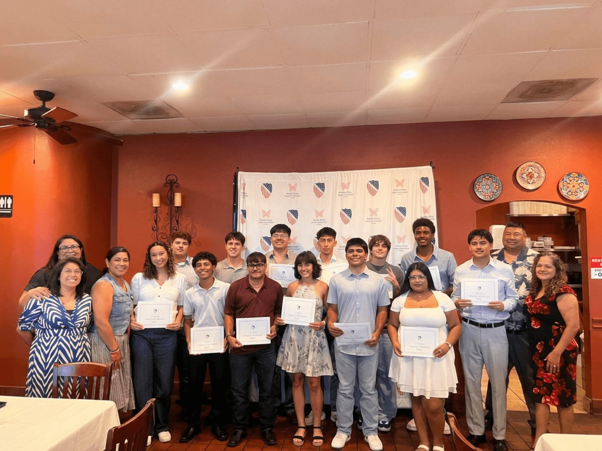 Group of people holding certificates in a restaurant with red walls and decorative plates, smiling for the photo.