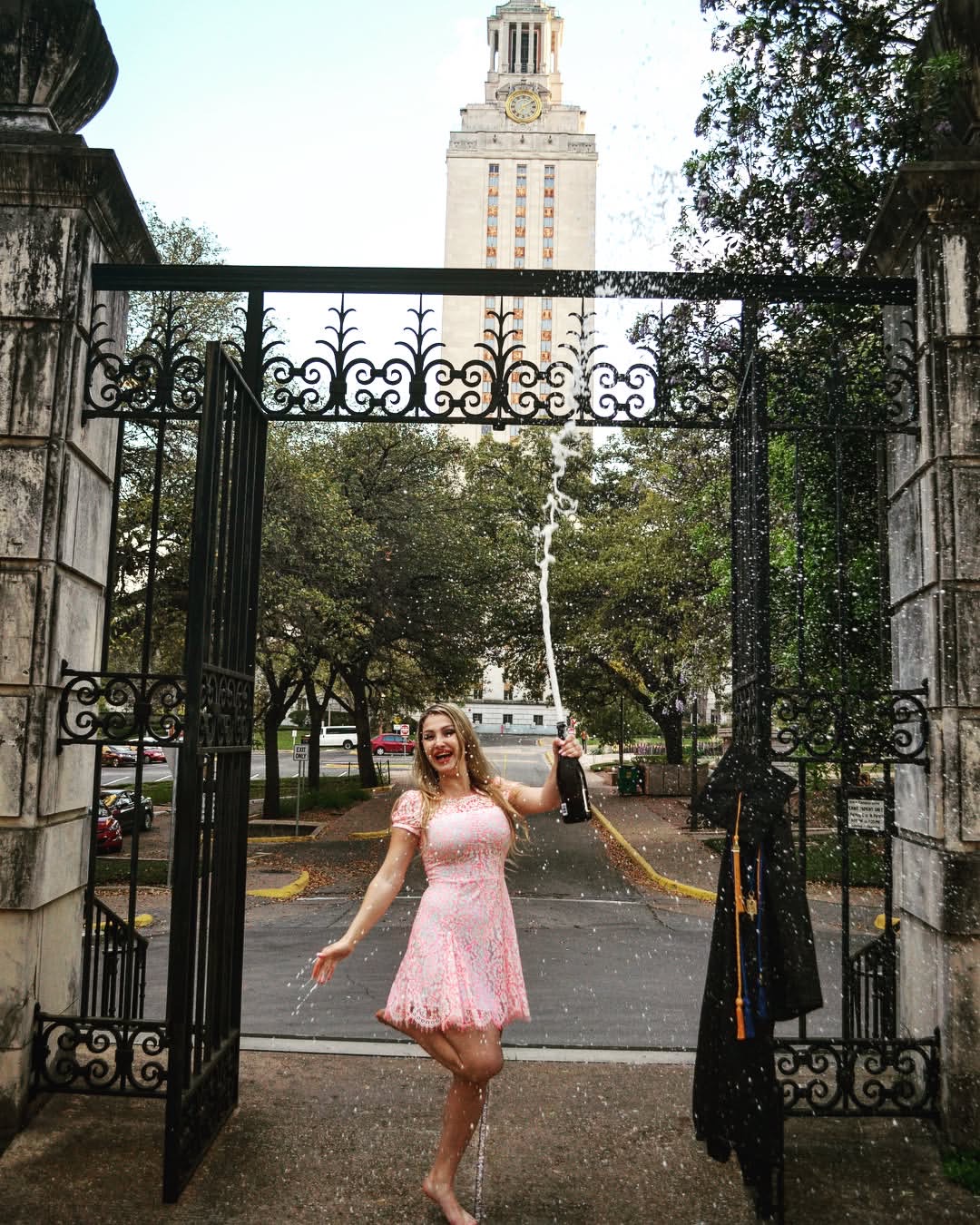Young woman in pink dress standing in open gate, holding a black item, with trees and a tall building in background.