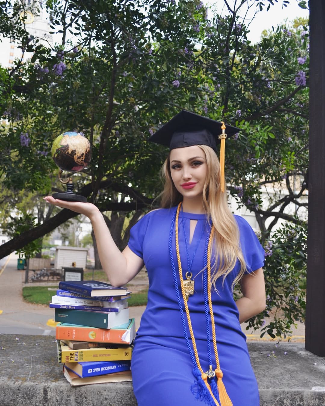 Young woman in graduation cap and gown holding a globe, standing outdoors with trees and books nearby.