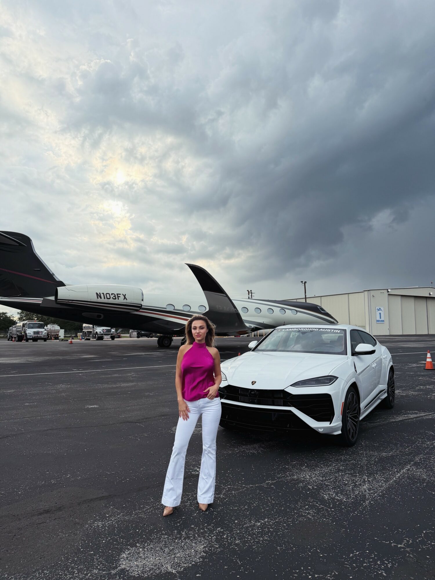 Woman standing in front of a white car and a private jet on an airport tarmac under cloudy sky.