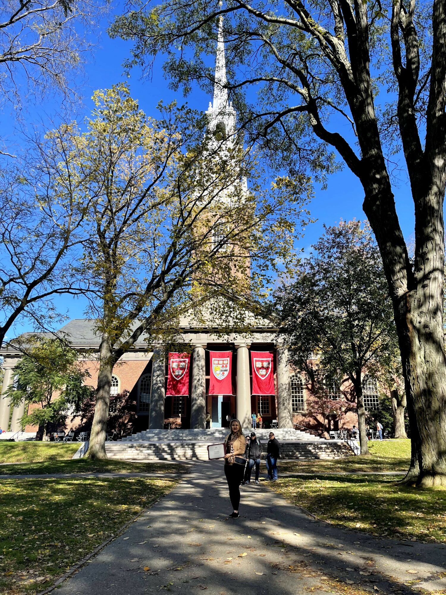 People walking on a path toward a building with banners, surrounded by trees and a clear blue sky.