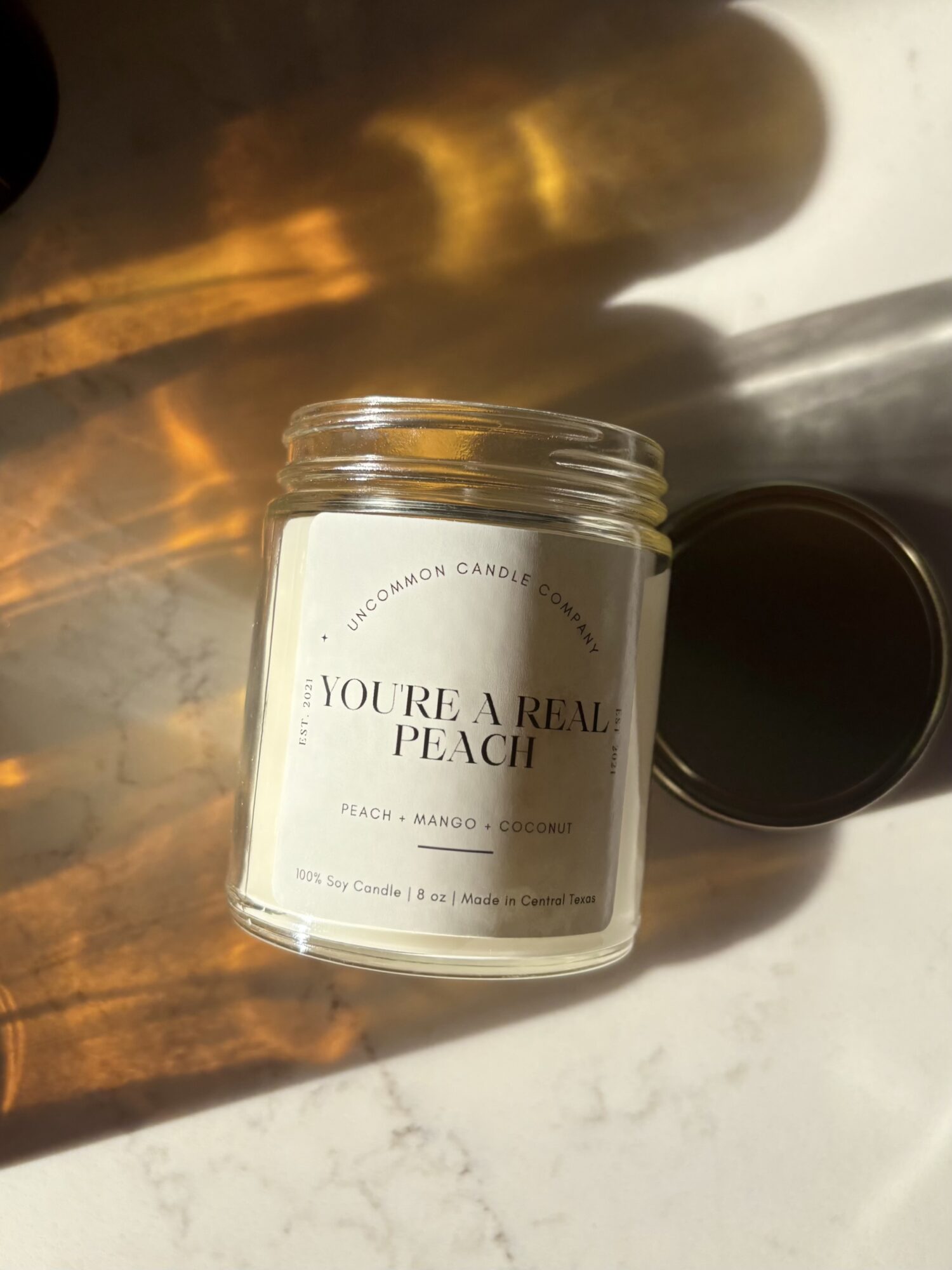 Open jar of peach-scented candle with black lid beside it, on a white surface with warm light reflection.