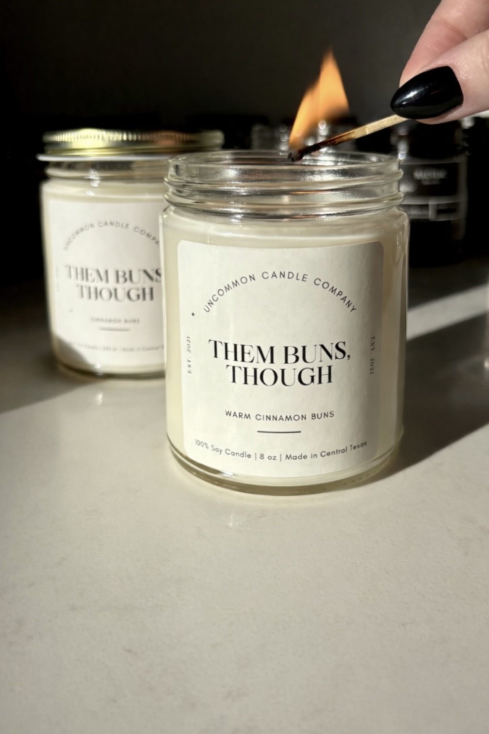 A lit match is held above a candle in a jar, with another jar candle in the background. The candles have white labels.