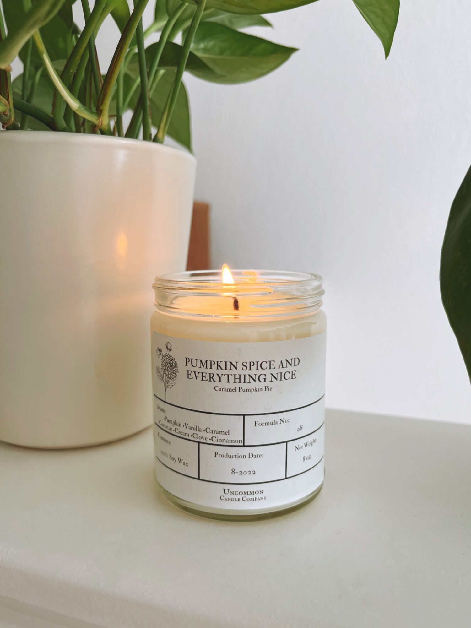 Lit candle in a glass jar with a label, placed on a white surface next to a potted plant with green leaves.