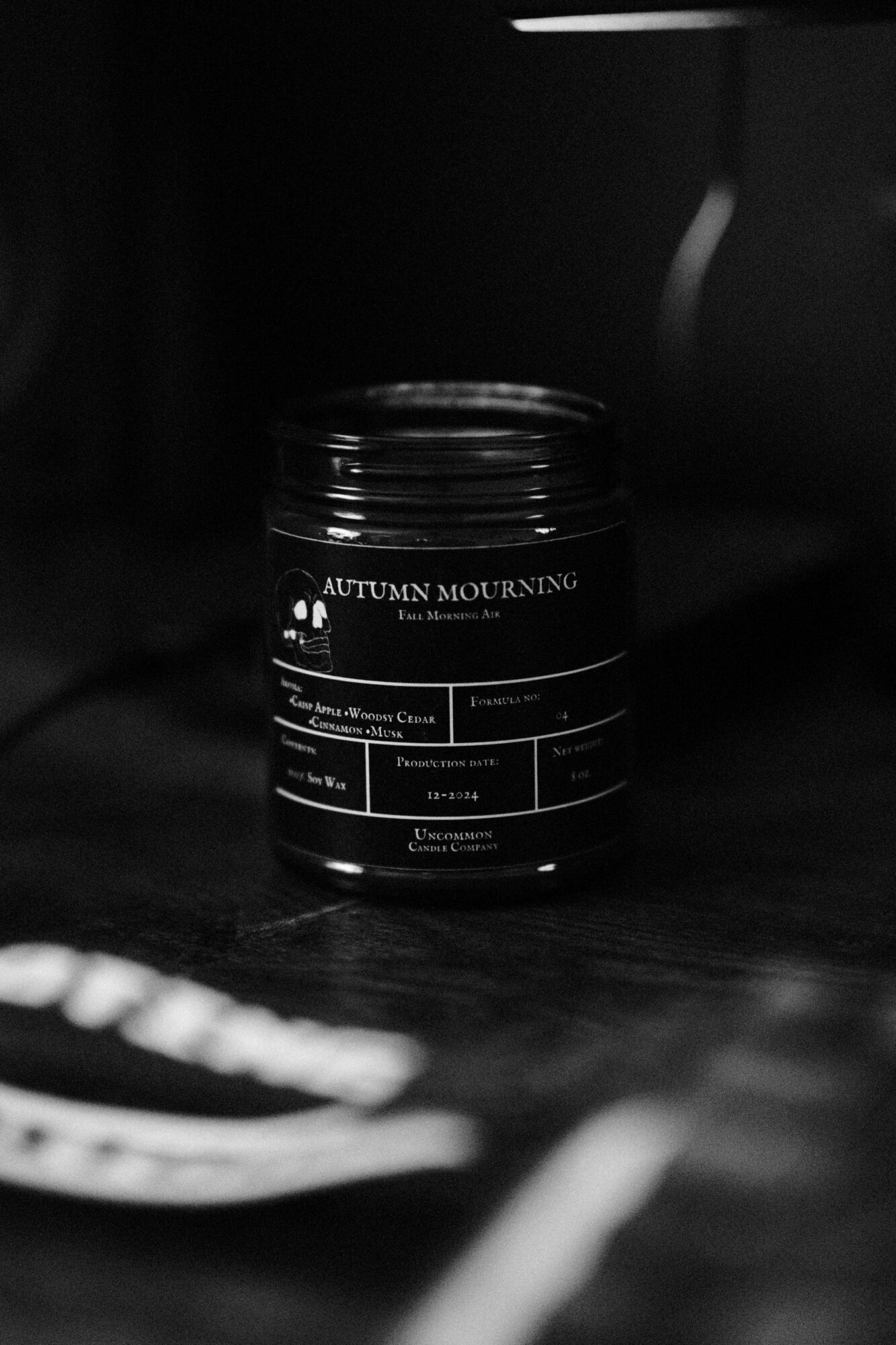 A black jar labeled 'Autumn Mourning' on a dark surface with a blurred object in the foreground.
