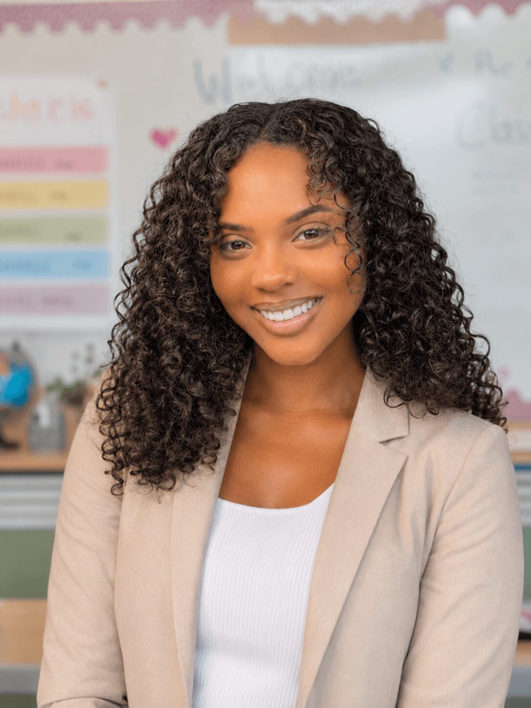 Smiling woman with curly hair in a classroom, wearing a beige blazer and white top.