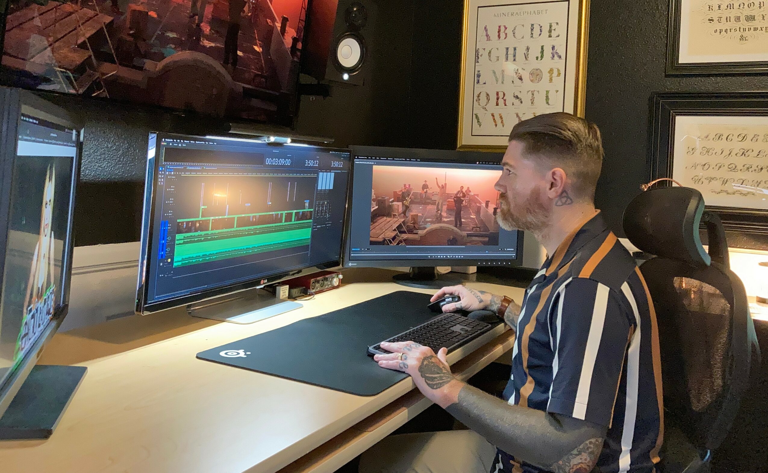 Person working at a desk with two monitors, editing video or audio, in a room with framed artwork on the wall.