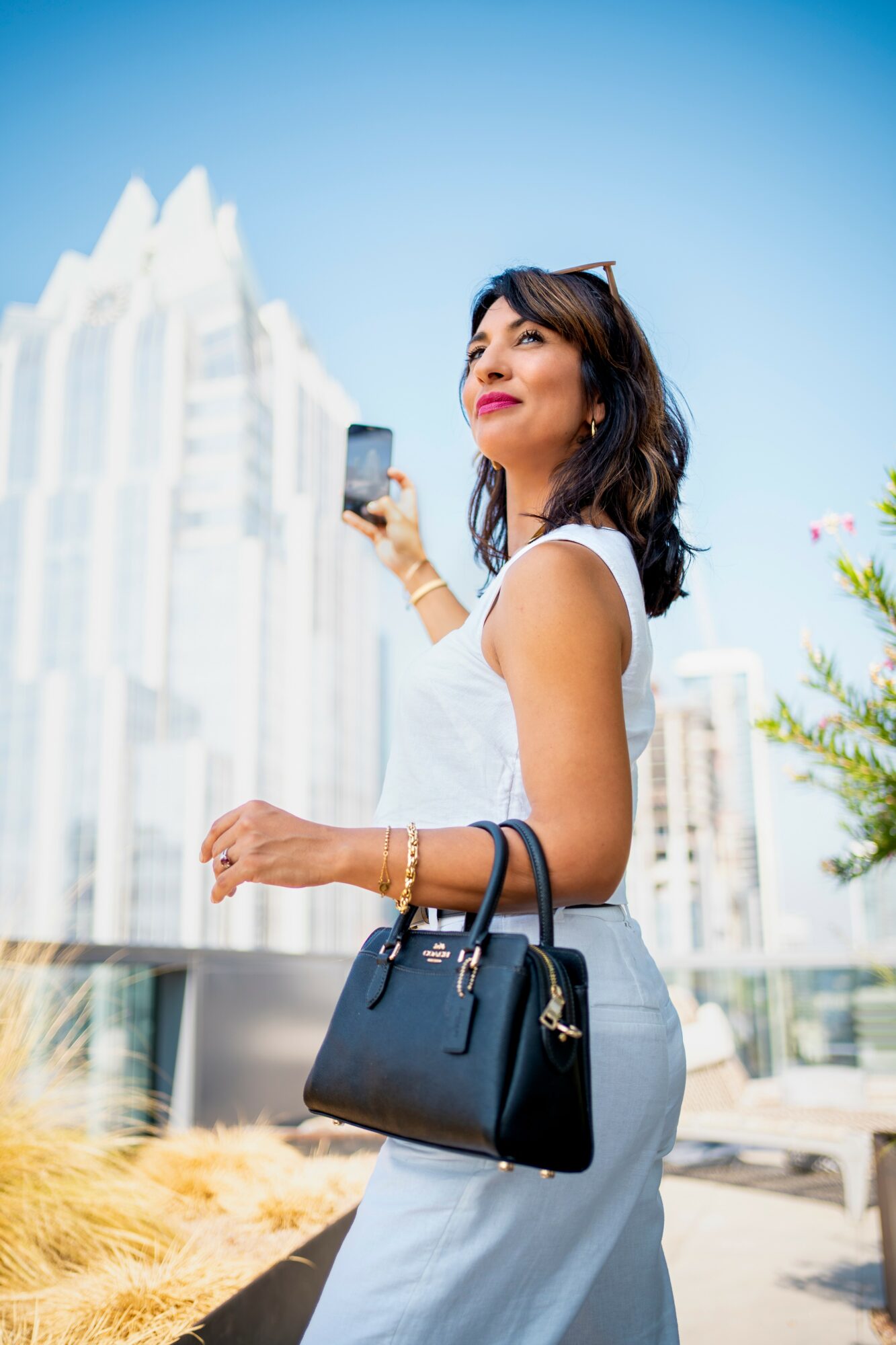 Woman in white dress holding a phone and a black handbag outdoors with city buildings in background.