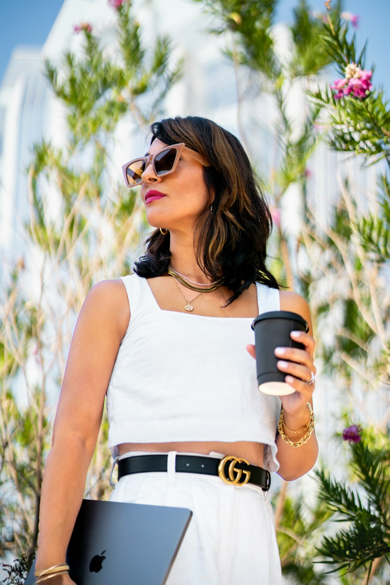 Woman in white dress with sunglasses holding a coffee cup outdoors, surrounded by tall green plants and pink flowers.