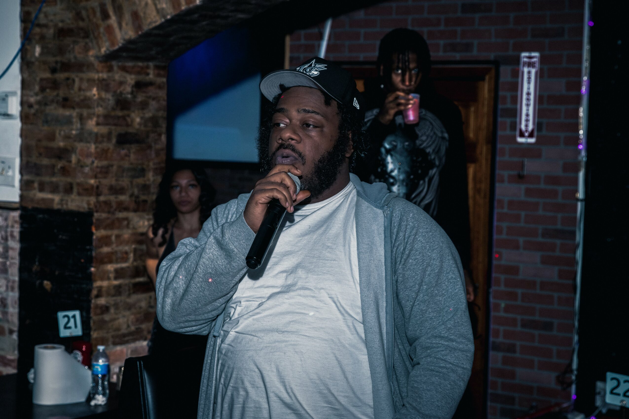 Man with beard and cap holding microphone, speaking at an indoor event with brick walls and people in background.