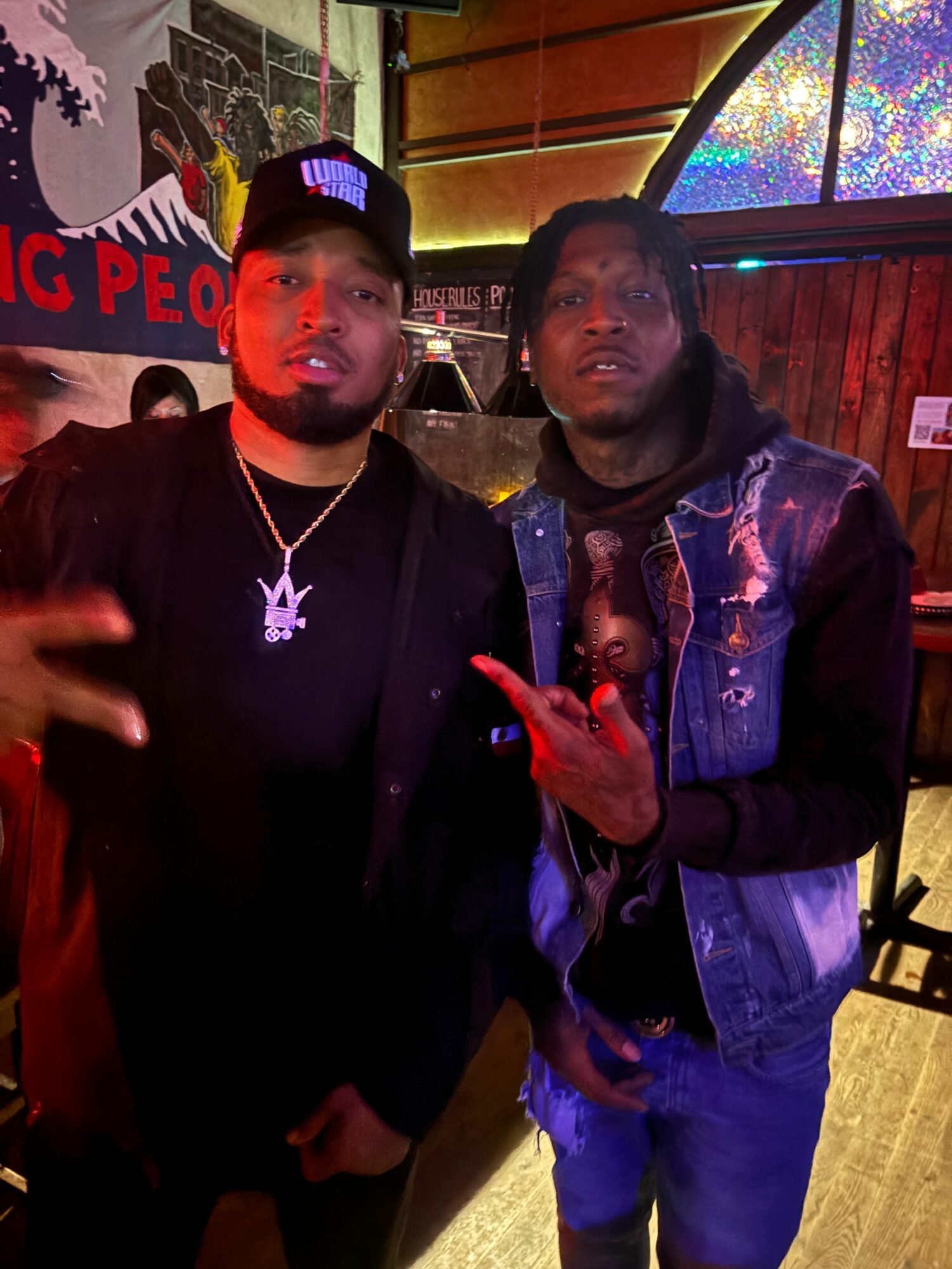 Two men standing indoors, one wearing a black hoodie with a necklace, the other in a denim jacket, both making hand gestures.