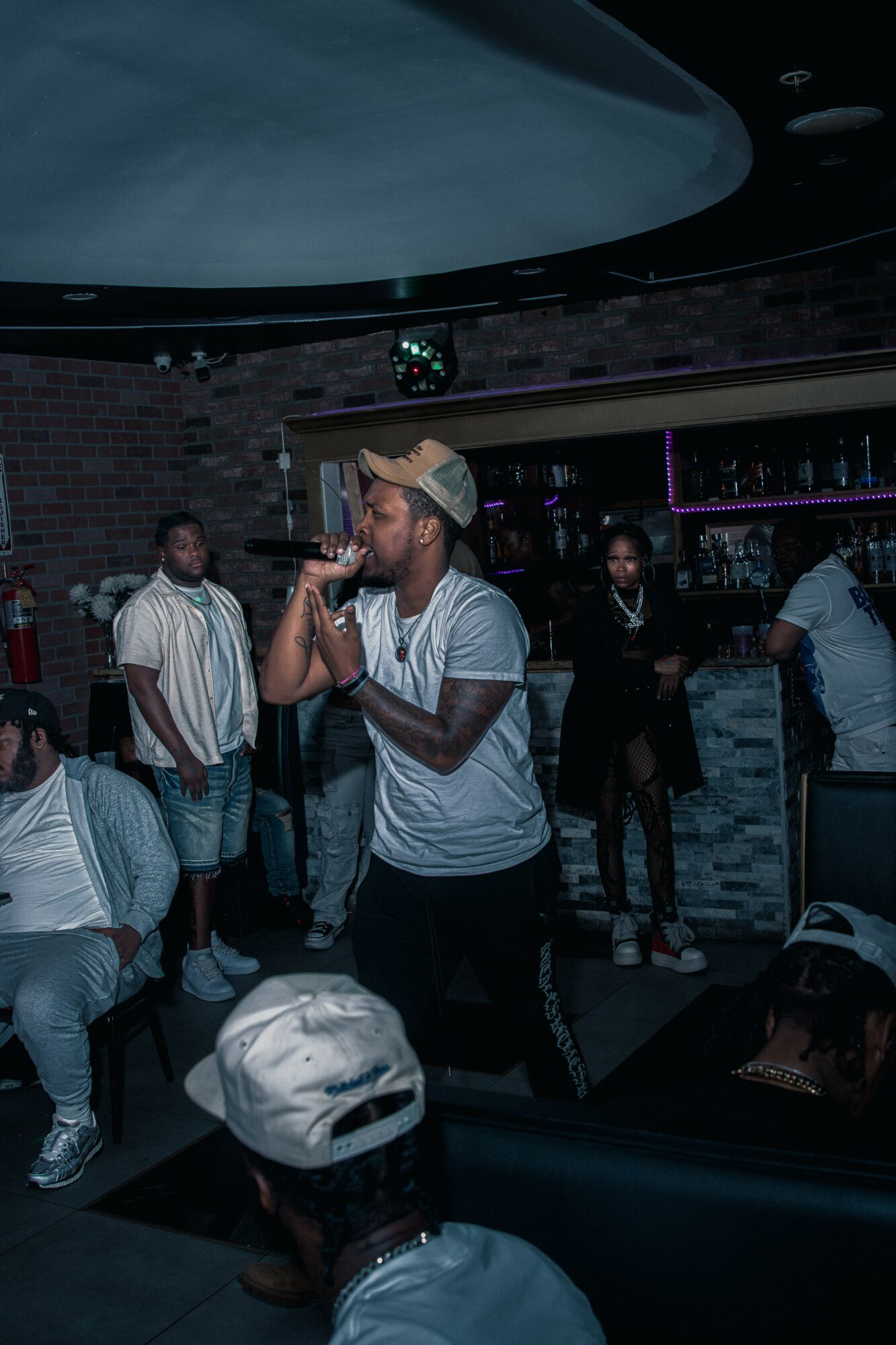 Person singing into a microphone in a dimly lit bar with other people in the background.