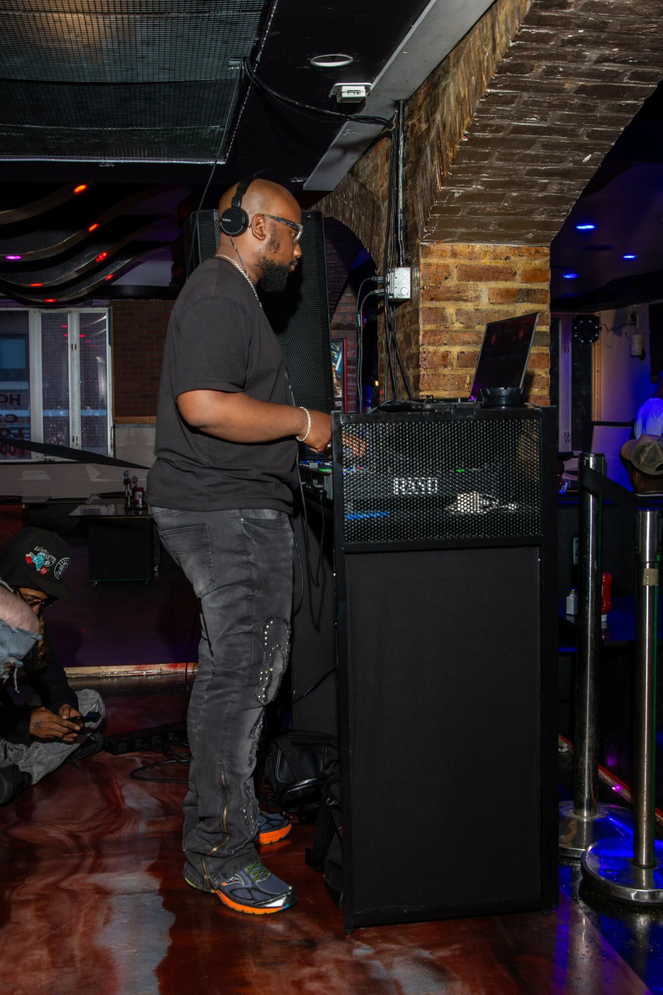 Man wearing headphones and sunglasses standing at DJ booth in a dimly lit venue with brick wall background.