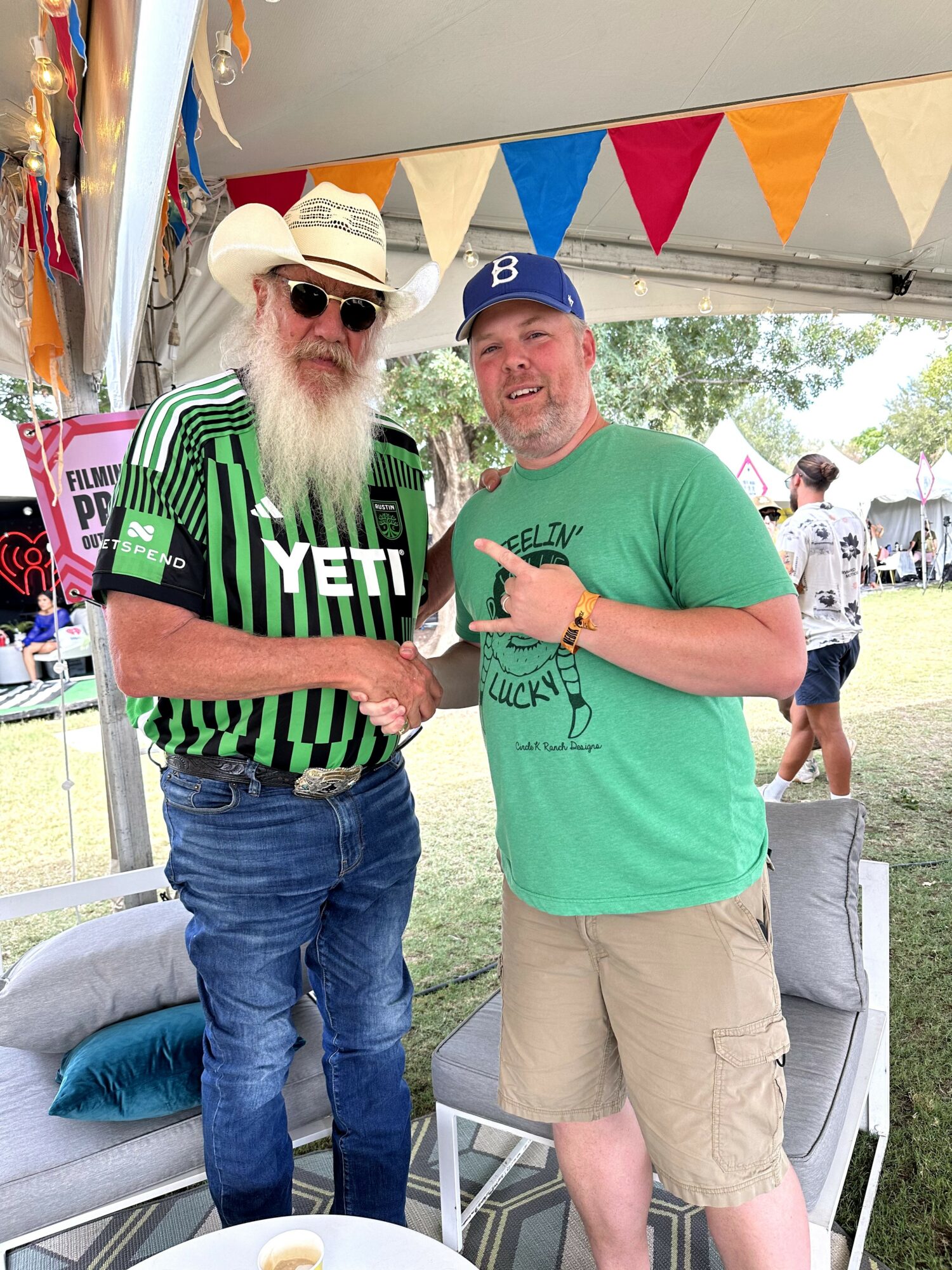 Two men shaking hands outdoors under a tent with colorful bunting, one with a long white beard and sunglasses, the other in a baseball cap.