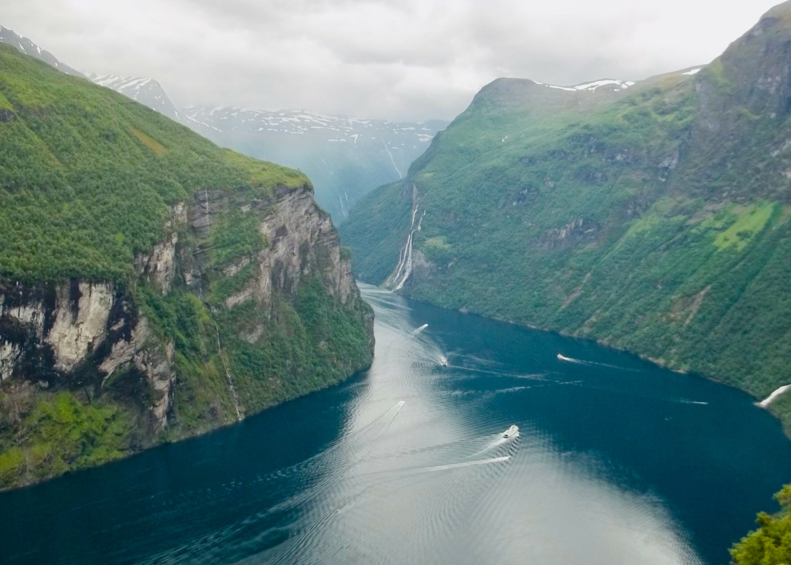 A fjord with steep green cliffs, water, and a waterfall in the distance under cloudy sky.