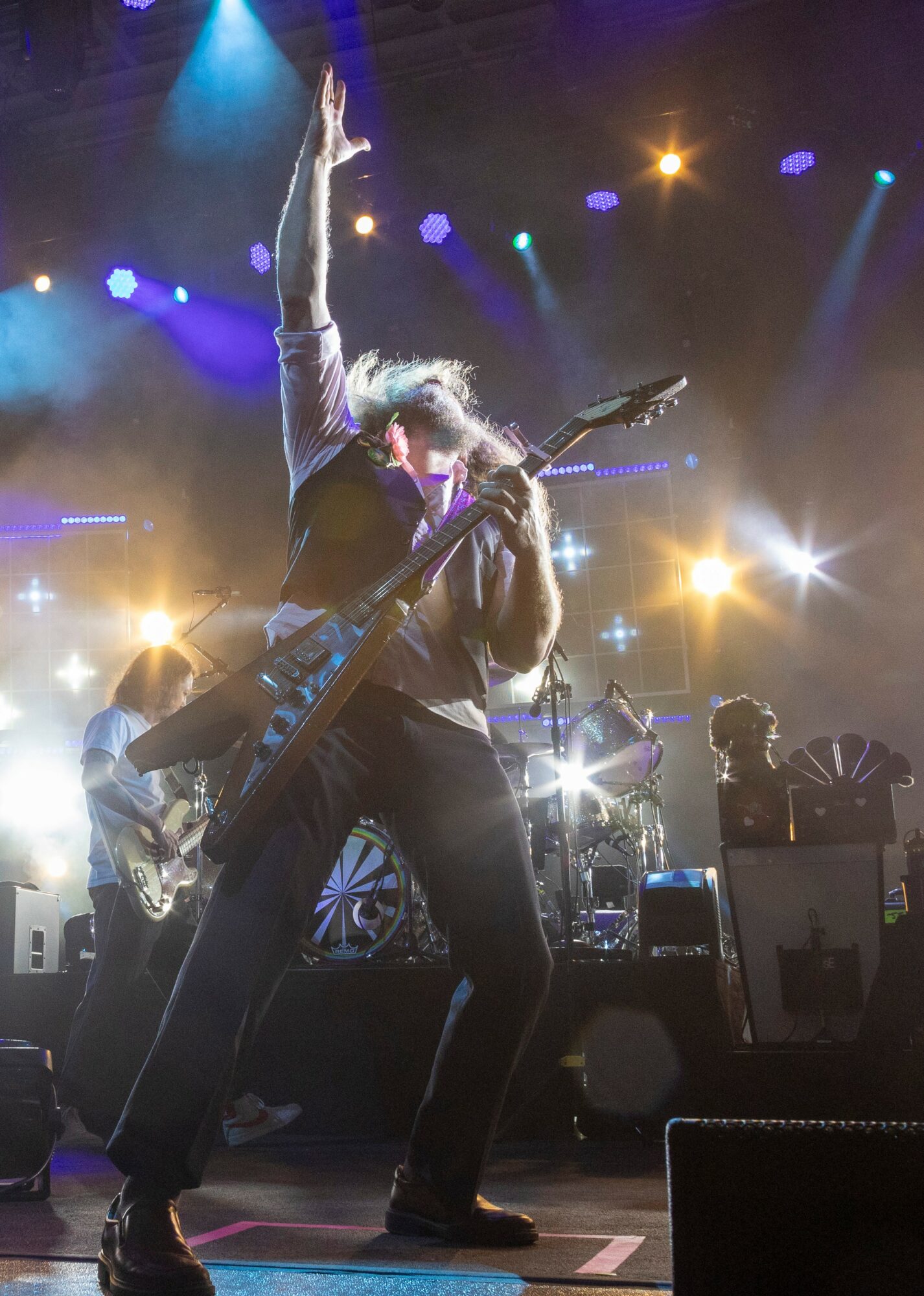 Musician on stage with guitar, raising one arm, surrounded by colorful stage lights and band members.