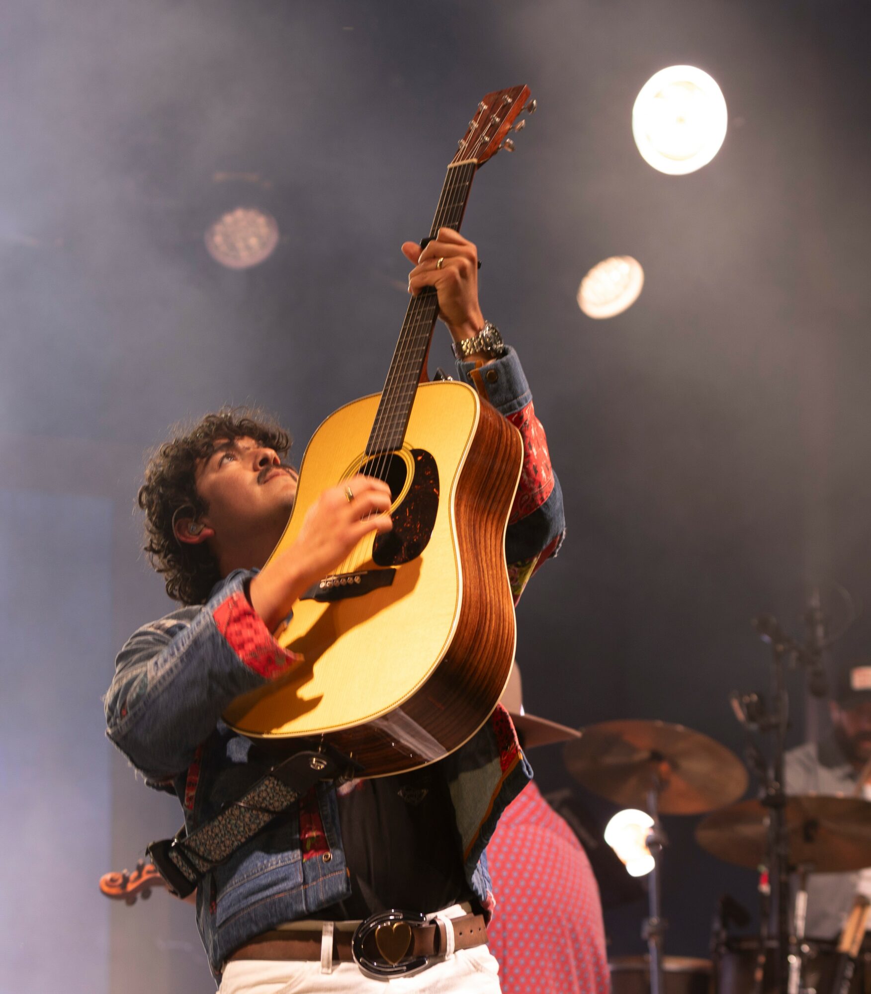 Child playing guitar on stage with bright lights and drums in background, looking up at the instrument.