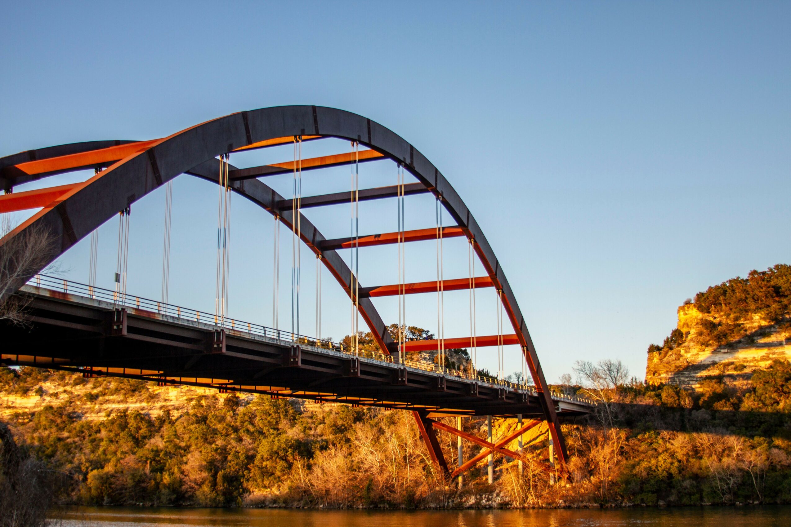 Bridge with red arches spans over a river, with trees and rocky cliffs in the background, under a clear blue sky.