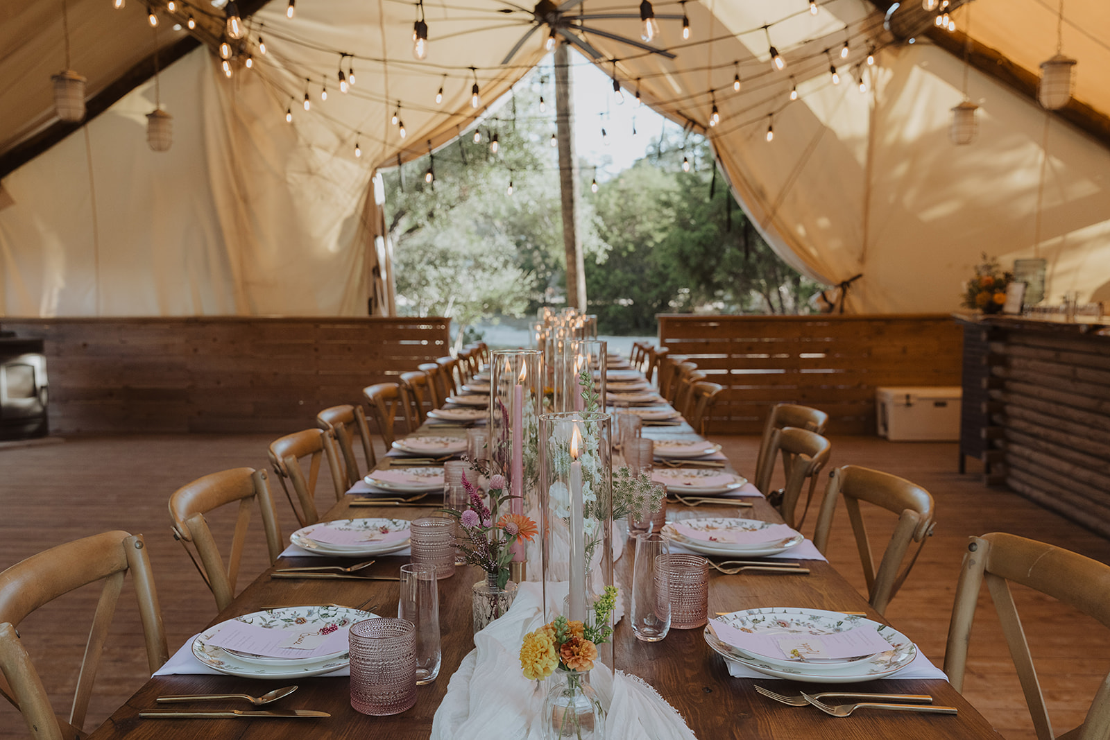 Long dining table set with plates, glasses, and floral centerpieces inside a tent with string lights and windows.