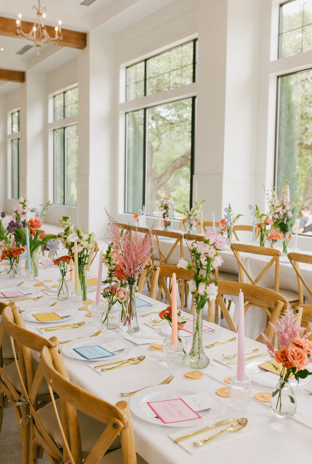 Long dining table with floral centerpieces, set with plates, glasses, and cutlery in a bright room with large windows.