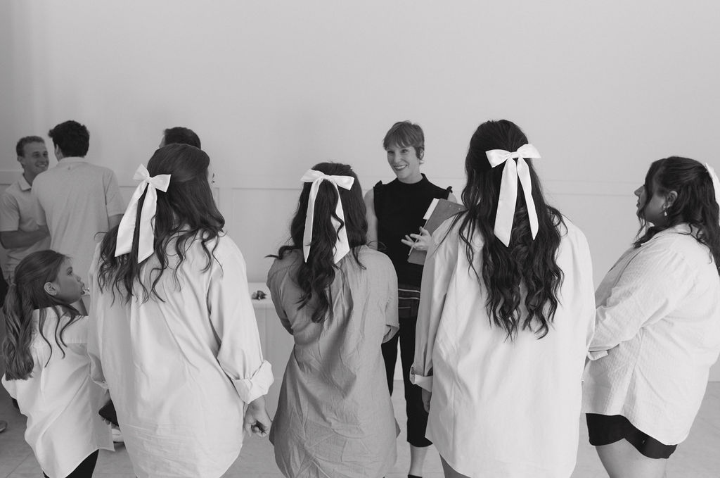 Group of children and a woman standing in a room, some children wearing white coats and ribbons, facing a woman speaking.