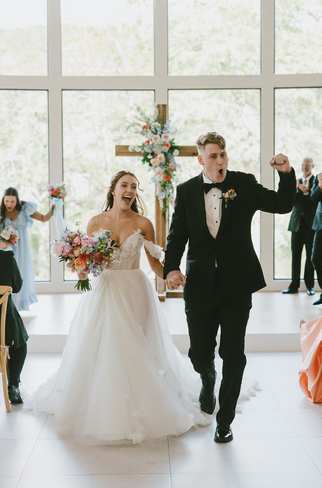 Bride and groom holding hands and smiling, walking in a bright wedding venue with large windows and floral decorations.